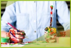 Zwei Kinder-Hände zu Fäusten geballt auf einem Blatt Papier. Die linke Hand hält einen Stift und die rechte Hand einen Pinsel in der Hand. Beide Hände und das Blatt Papier sind voll mit bunter Farbe.