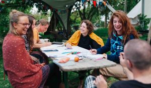 Fünf Frauen sitzen an einem Tisch im Garten, auf dem ein großes weißes Blatt Papier mit Stiften liegen. An der Stirnseite am rechten unteren Bildrand sitzt noch eine Person mit ganz kurzen Haaren mit Blick zu den Frauen.
